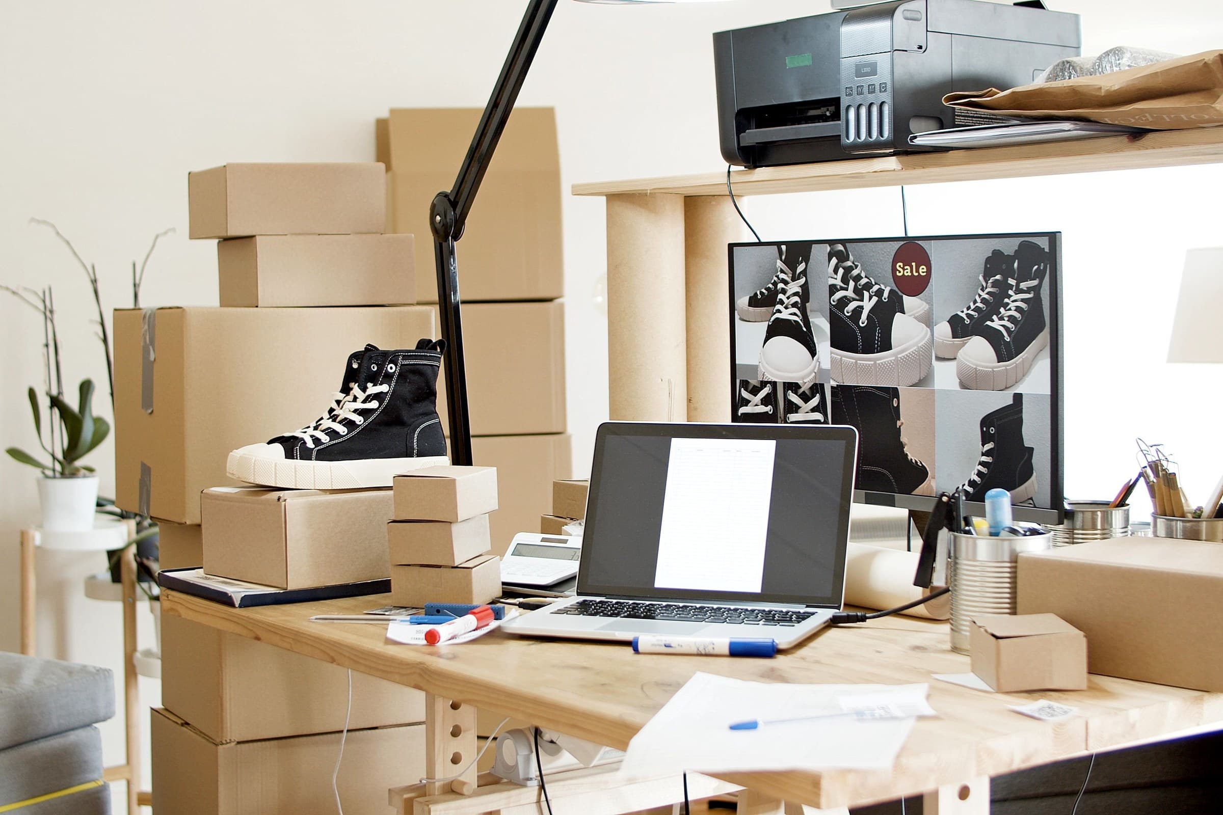 An e-commerce packing desk with shipping cartons, a laptop and a storefront on screen.